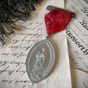 May include: An oval silver-toned metal religious medal featuring a relief of a figure in robes, with the Latin text COR JESU SACRATISSIMUM MISERERE NOBIS. The medal is attached to a faded red fabric ribbon with a metal pin clasp at the top.