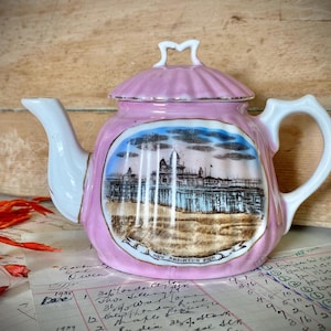 Könnte beinhalten: Eine rosa Keramikteekanne mit geriffeltem Deckel sowie weißem Henkel und Ausguss. In der Mitte befindet sich eine detaillierte Abbildung des New Brighton Pier mit blauem Himmel und braunem Vordergrund, beschriftet mit New Brighton Pier.
