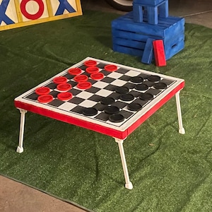 May include: A red and white checkerboard game table with black and red checkers. The table has white legs and is set up on a green artificial grass surface.