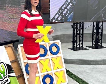 May include: A woman in a red and white striped dress holds a yellow X game piece in front of a large wooden tic-tac-toe game. The game board has blue O's and yellow X's.
