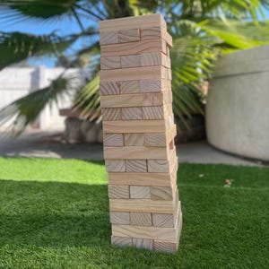 May include: A tall tower of wooden blocks stacked in a game of giant Jenga. The blocks are natural wood color and are stacked in a staggered pattern. The tower is standing on a green artificial grass surface.