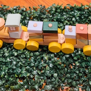 May include: A wooden toy train with colorful blocks. The train has a green, blue, and pink engine with yellow wheels. The blocks are stacked on top of each other and are in various colors, including green, blue, pink, and brown.