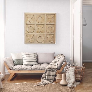 May include: A light gray living room with a wooden futon couch, a white throw blanket, and a wooden wall art piece with the letters "XO" repeated in a grid pattern.