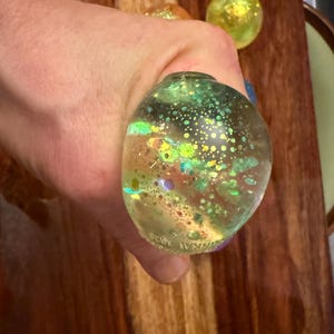 May include: A clear, oval-shaped stress ball filled with iridescent glitter and small bubbles. The ball is held in a hand, with two other glitter-filled spheres visible in the background. The ball has a green and yellow color scheme.