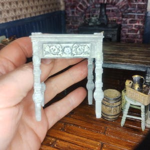May include: A miniature, light gray table with ornate floral carvings on the front. The table has four turned legs and is designed for a dollhouse or miniature scene. The table is held in a hand.