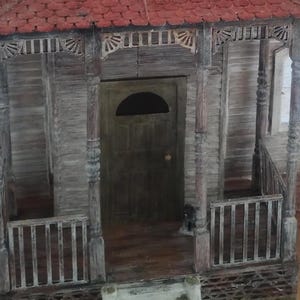 Può includere: Una casa in miniatura di legno con tetto rosso e portico. La casa ha una porta in legno scuro con una finestra a semicerchio. Il portico ha finiture decorative e una piccola ringhiera. Una piccola figura nera è sul portico.