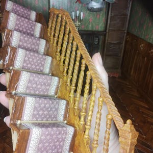Può includere: Una scala in legno in miniatura con finitura marrone e dorata. La scala ha un tappeto a motivi e un corrimano in corda intrecciata.