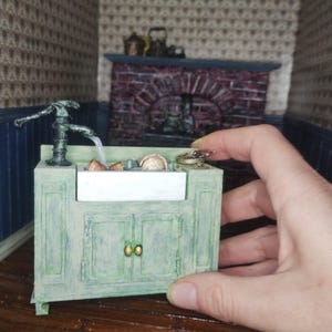 May include: A miniature green painted cabinet with a white porcelain sink and a copper faucet. The cabinet has two doors with gold knobs. The sink is filled with miniature dishes and a small copper pot.