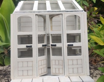 May include: A white wooden miniature greenhouse with a glass roof and open doors. The greenhouse has a small wooden porch with a rounded edge.
