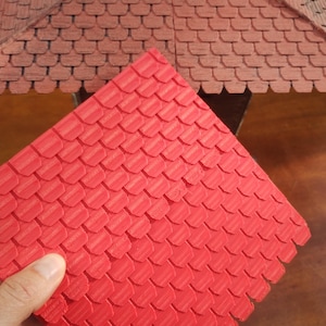 May include: A red, textured, scale-like roof tile held in hand, with a matching roof structure in the background. The tiles are a vibrant red, and the background roof is a similar design in a muted reddish-brown colour.