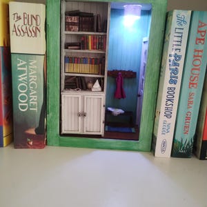 May include: A miniature bookshelf diorama with a light inside. The bookshelf is filled with miniature books and has a small door with two knobs. The diorama is set inside a green frame and is sitting on a shelf with other books.