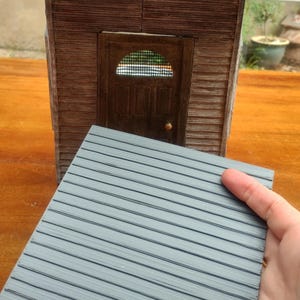 May include: A miniature wooden house with a brown door and a gray, ridged panel. The door has a small, arched window with a mesh screen. The house's exterior is made of textured wood panels.
