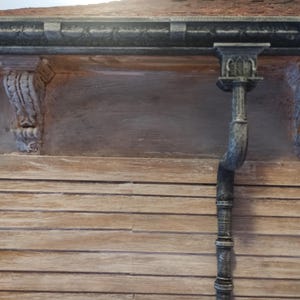 Peut inclure: Gros plan sur l'extérieur d'un bâtiment, mettant en valeur des détails architecturaux. L'image présente une façade en bois avec des corbeaux décoratifs et une descente pluviale. La palette de couleurs est dominée par des bruns et des gris sourds, donnant un aspect vintage.