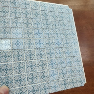 May include: A square sheet of miniature tiles featuring a repeating blue and white floral pattern. The grid consists of small, individual square tiles arranged in rows and columns, displaying a detailed, symmetrical decorative design on each unit.