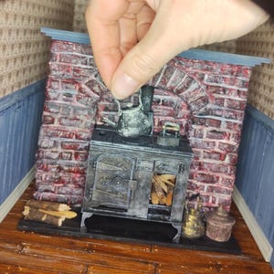 May include: A miniature dollhouse kitchen scene featuring a black cast iron stove, a brick fireplace, and a metal kettle. The stove has a door with a metal handle and a window revealing logs inside. The kettle is positioned on top of the stove.