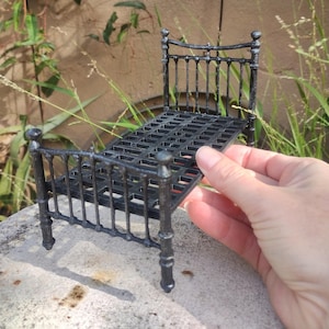 May include: A miniature black metal bed frame with a headboard and footboard. The bed frame has a slatted base and is designed with vertical bars. The bed frame is made of metal and is designed for a dollhouse or miniature setting.