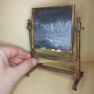 May include: Miniature wooden chalkboard with a dark gray surface and a brown frame. The chalkboard is mounted on a wooden stand. A small piece of chalk and a green eraser are on the ledge. The chalkboard has some white markings on it.