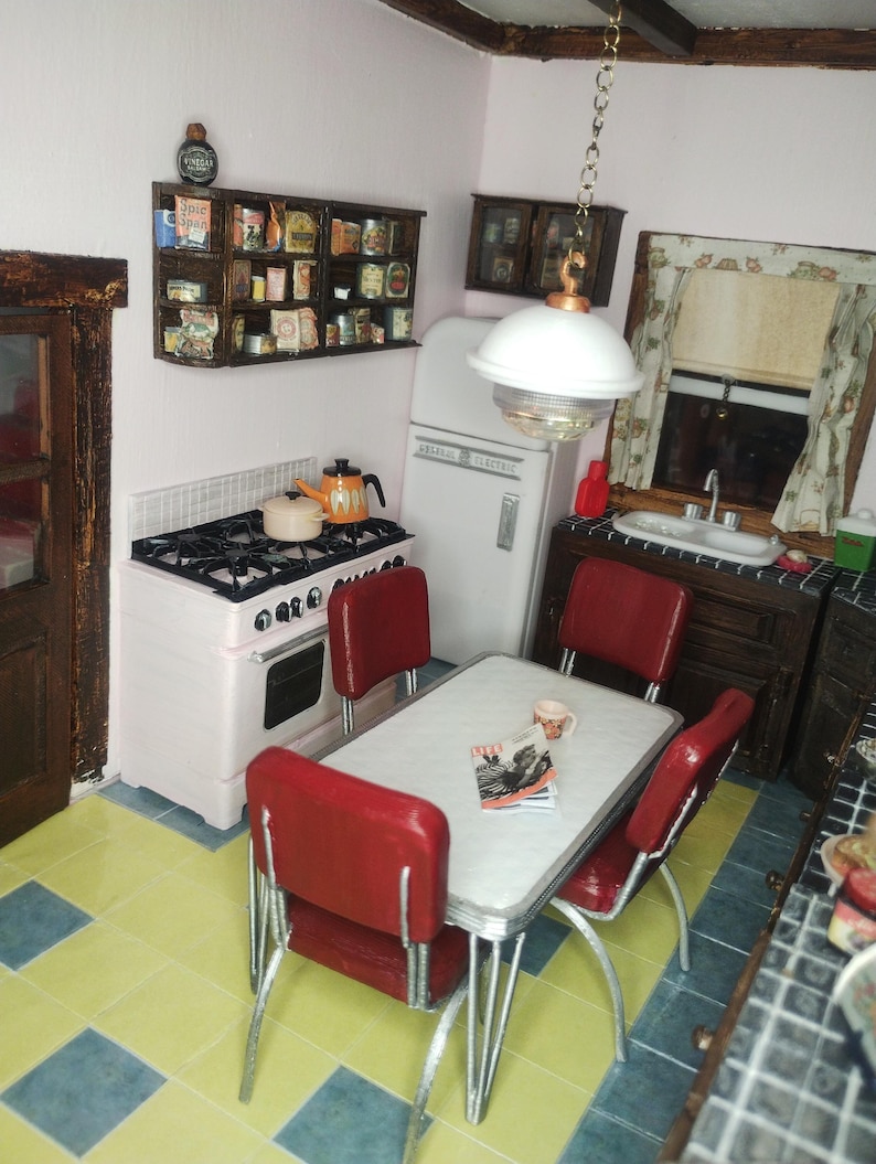Pu&ograve; includere: Scena di cucina retr&ograve; in miniatura con una stufa rosa e bianca, un frigorifero bianco e un tavolo con sedie rosse. Una mensola espone cibi in miniatura. Un lampadario illumina la scena.