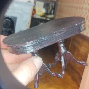 May include: A miniature, dark-colored table with an oval top and ornate legs. The table appears to be made of wood and is painted with a dark finish. The legs have a detailed, decorative design. The table is approximately 6 inches long.