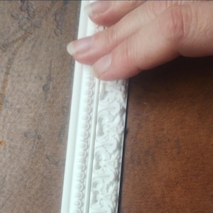 May include: A white decorative molding strip featuring a raised relief pattern. The molding consists of two distinct sections: one side displays a repeating bead-like circular design, while the other side features an intricate floral scrollwork.