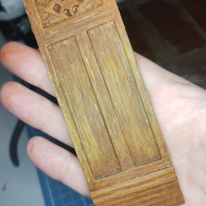 May include: A miniature wooden door with a detailed design. The door is painted in shades of brown and yellow, with a decorative top panel and two vertical panels below. The wood grain is visible, adding texture. The door is likely for a dollhouse or model.