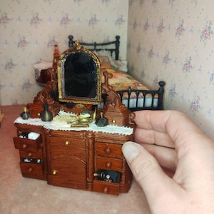 May include: Miniature wooden vanity unit with a mirror, drawers, and decorative items. The vanity is brown with gold accents and a white lace trim. The mirror has a gold frame. The drawers are open, revealing small items inside. Part of a dollhouse bedroom set.