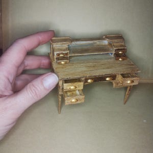 May include: Miniature wooden desk with a light brown finish and gold-colored drawer pulls. The desk has several small drawers and a hutch-like top. The desk is held by a hand, showcasing its small size, perfect for a dollhouse or craft project.