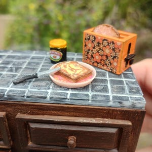 May include: Miniature kitchen scene with a slice of toast on a plate, a jar of Marmite, and a floral-patterned toaster. The toast is buttered. The toaster is orange with a black lever. The countertop is gray and white tile.