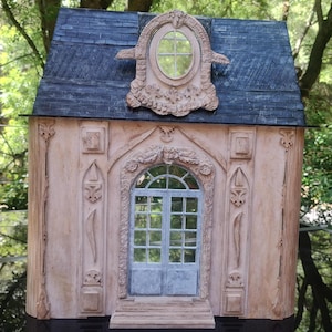 Puede incluir: Una fachada de casa de muñecas en miniatura con un diseño detallado y ornamentado. El frente de la casa está pintado de color rosa claro con molduras blancas. El techo es de pizarra gris oscuro. La casa tiene una gran puerta arqueada con una puerta doble y una ventana redonda más pequeña encima. La casa está decorada con intrincados tallados y molduras.