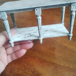 May include: A miniature, distressed-look table with a rectangular top and a lower shelf. The table is painted in shades of white and gray, with ornate detailing on the legs and corners. The table is approximately 6 inches long.