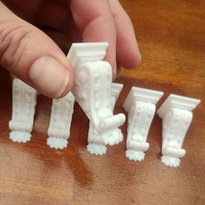 May include: A hand holding several small, white decorative corbels. The corbels feature an ornate design with scrollwork and floral patterns. They are made of a solid, opaque material.