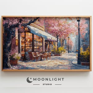 May include: A framed painting depicts a charming street scene with a cafe, cherry blossom trees, and a lamppost. The cafe has a white awning and inviting interior lighting. The artwork is in a wooden frame.