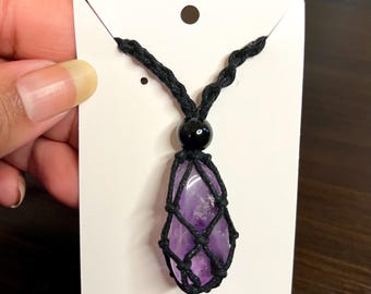 Puede incluir: Un collar con colgante de amatista púrpura. La amatista está encerrada en una red negra tejida, con una cuenta negra encima. El collar está en una tarjeta blanca con un agujero en la parte superior.