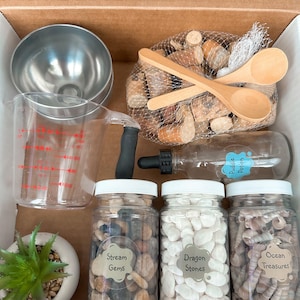 May include: A cardboard box filled with sensory play items. Includes a metal bucket, measuring cup, wooden spoons, a net bag of wooden pieces, a bottle labeled "Puddle Potion", jars of stones and shells, and a small succulent.