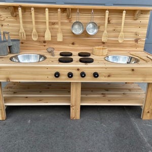 Könnte beinhalten: Eine hölzerne Outdoor-Spielküche mit zwei Edelstahlspülen, Holzutensilien und Kochtöpfen. Die Spielküche hat eine hölzerne Rückwand mit hängenden Utensilien und ein unteres Regal zur Aufbewahrung. Die Spielküche ist aus hellem Holz gefertigt.