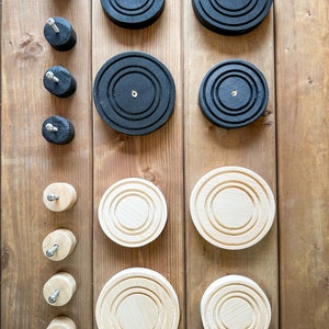 May include: Overhead view of wooden objects. Black, circular coasters with concentric grooves and gold-coloured centre accents are displayed. Natural wood coasters and matching wooden pegs with metal screws are also visible, all arranged on a wooden surface.