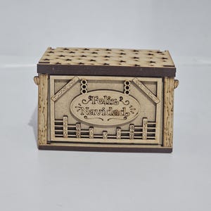 May include: A rectangular wooden box with a dark brown lid featuring a laser-cut star pattern. The front panel displays an oval plaque engraved with the text Feliz Navidad, surrounded by decorative fence-like details and wood grain textures.