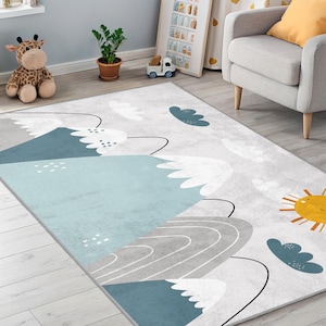 Puede incluir: Una alfombra rectangular con un diseño de montaña y sol. La alfombra presenta una combinación de colores azul claro, gris y blanco. El diseño incluye montañas estilizadas, nubes y un sol amarillo. La alfombra está colocada sobre un suelo de madera clara.