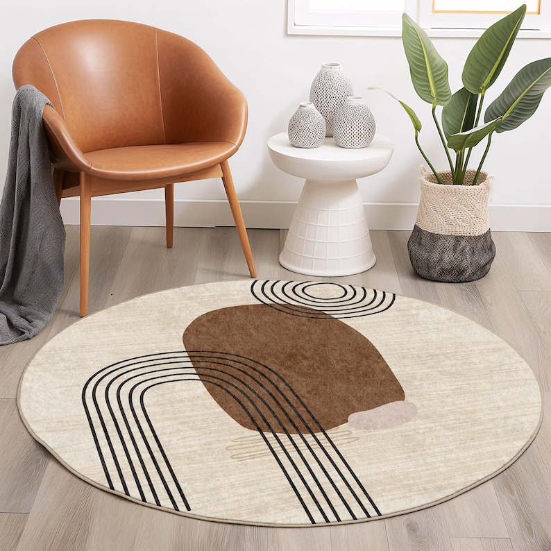 May include: A round, cream-colored rug with a brown and black abstract design. The design features a large brown shape with black lines that resemble arches. The rug is on a wooden floor.