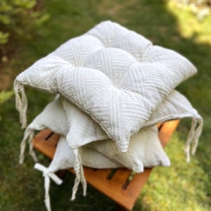 Può includere: Una pila di quattro cuscini quadrati color bianco sporco con motivi geometrici strutturati. Ogni cuscino ha un design trapuntato e lacci in tessuto attaccati. I cuscini sono impilati su un piccolo sgabello di legno, con uno sfondo verde sfocato.