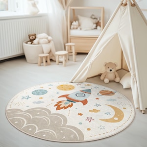 Puede incluir: Una alfombra redonda de color crema con un diseño de cohete, estrellas, planetas y una luna creciente. La alfombra es perfecta para la sala de juegos o el dormitorio de un niño.