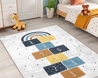 Puede incluir: Alfombra rectangular para niños con diseño de rayuela. La alfombra presenta un arcoíris y cuadrados numerados en tonos de azul, amarillo y marrón sobre un fondo blanco. Puntos negros dispersos.