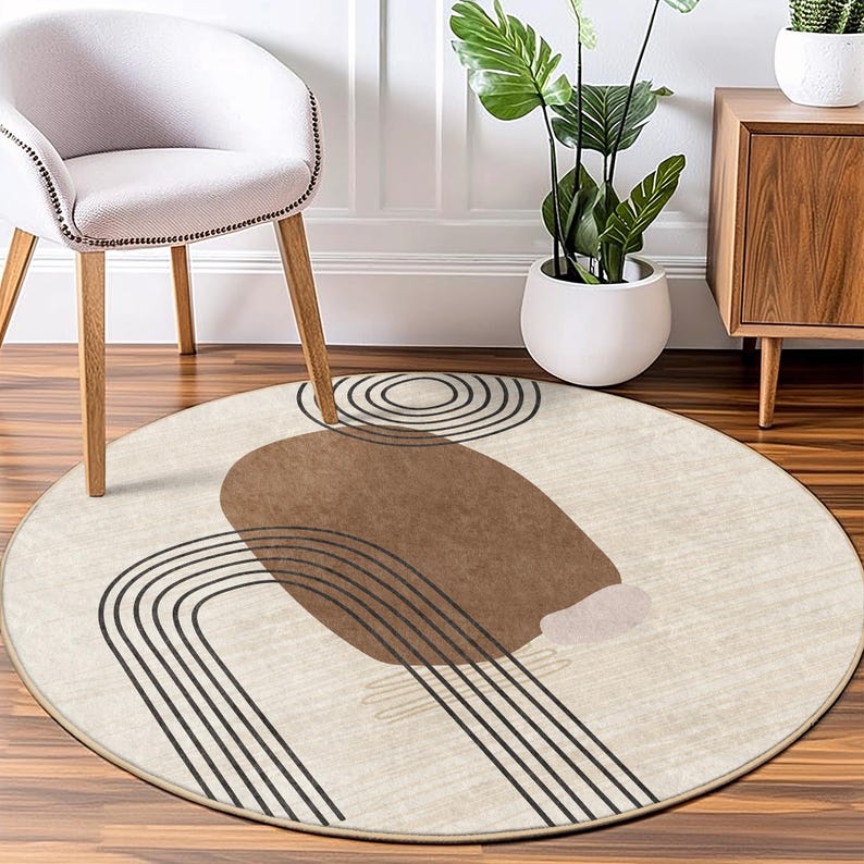 May include: A round beige rug with a brown abstract design featuring curved lines and a large brown circle. The rug has a subtle texture and a fringe border.