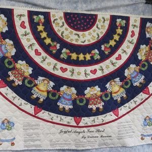 May include: A quilted Christmas tree skirt with a navy blue and white design. The skirt features a circular pattern with angels, stars, hearts, and holly. The text "Joyful Angels Tree Skirt" is visible.