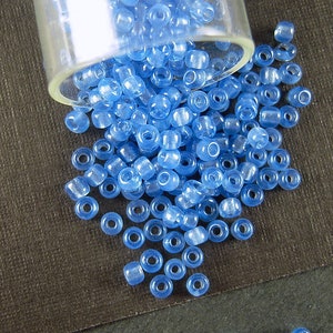 May include: A glass jar filled with light blue seed beads. The beads are scattered on a dark brown surface.