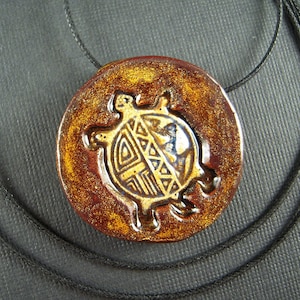 May include: A round, ceramic pendant with a turtle design. The pendant is brown with a gold turtle design in the center. The turtle design has geometric patterns. The pendant hangs on a black cord.