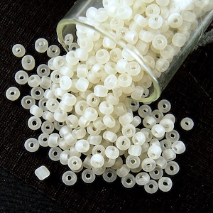 May include: A glass jar filled with white opaque seed beads, spilling out onto a black surface. The beads are round with a small hole in the center.