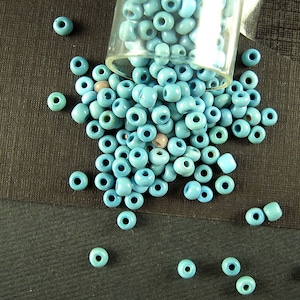 May include: A glass jar filled with light blue seed beads, spilling out onto a dark gray surface. The beads are small and round, with a smooth, glossy finish.
