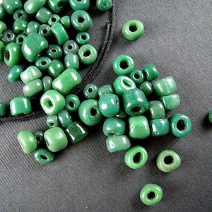 May include: A large pile of green glass beads, some are round and some are cylindrical. The beads are scattered on a dark gray surface.
