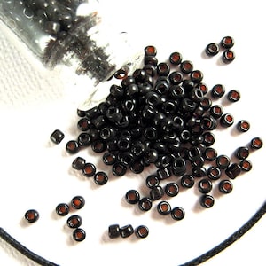 May include: A small glass jar filled with black seed beads with a brown center. The jar is tipped over and the beads are spilling out onto a white surface.